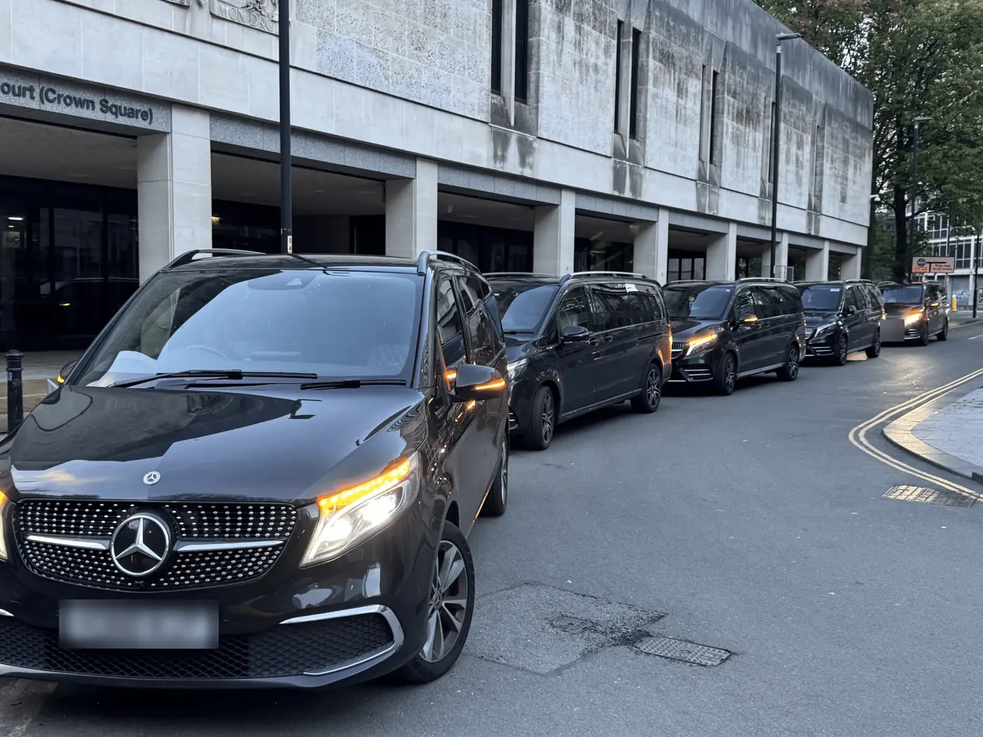 A line of black Mercedes V-Class chauffeur cars parked outside a building on Crown Square in Manchester