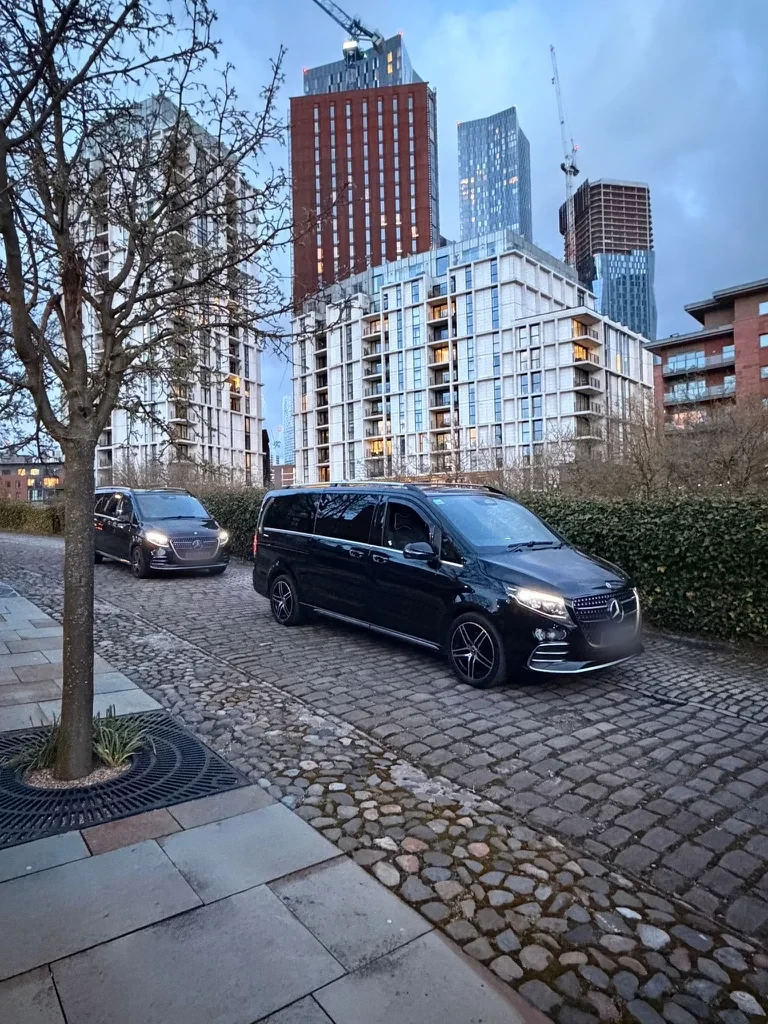 Two black Mercedes-Benz V-Class chauffeur cars parked on a cobbled street in Manchester at dusk, with apartment towers behind