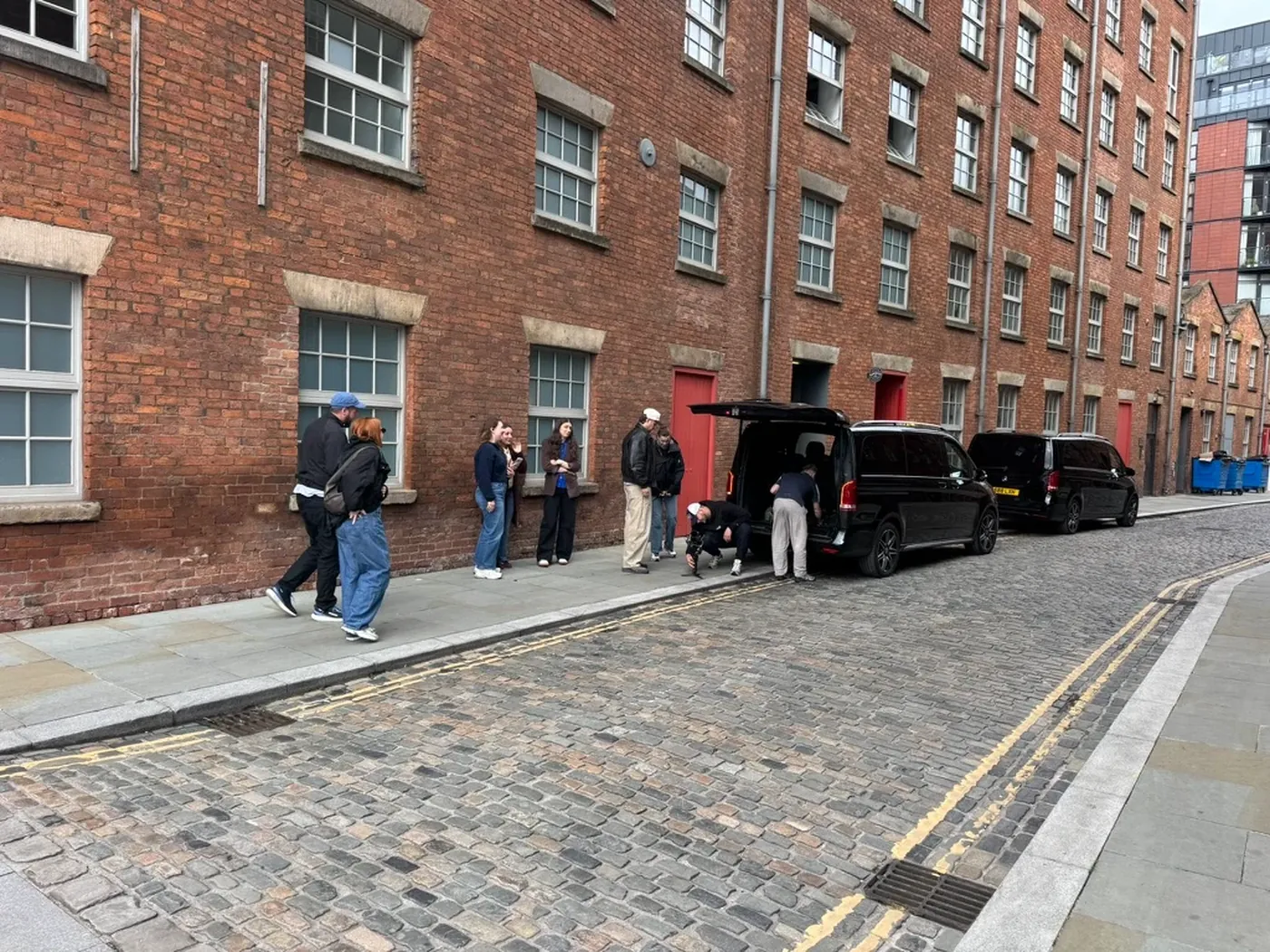 Two black Mercedes-Benz V-Class cars unloading a film crew on a cobbled Manchester street