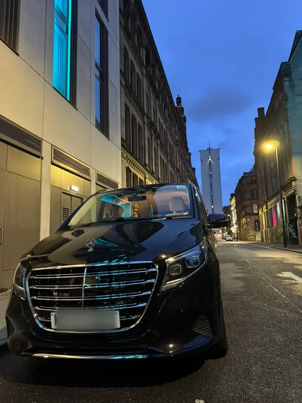 A black Mercedes V-Class chauffeur car on a Manchester city-centre street at night with a skyscraper lit up in the distance