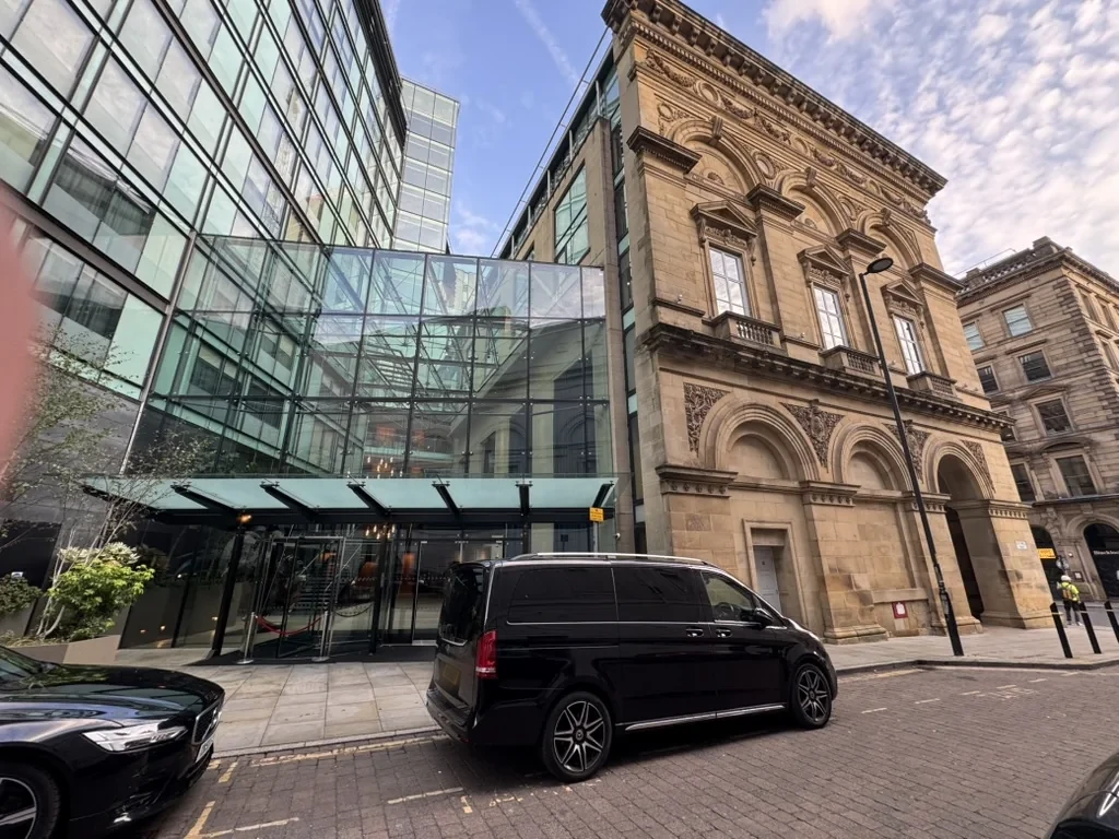 Black Mercedes-Benz V-Class chauffeur car outside a glass-fronted hotel entrance in central Manchester